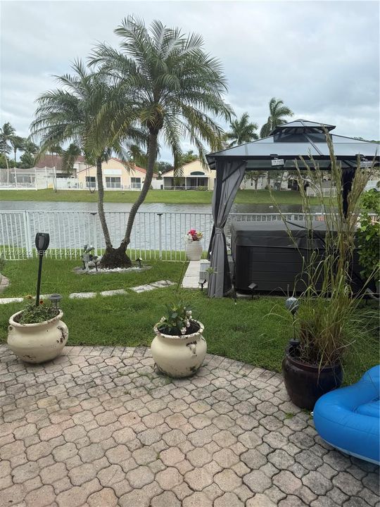 Backyard featuring a paved stone patio, permanent metal-frame gazebo, waterfront position with perimeter fencing, and mature palm trees