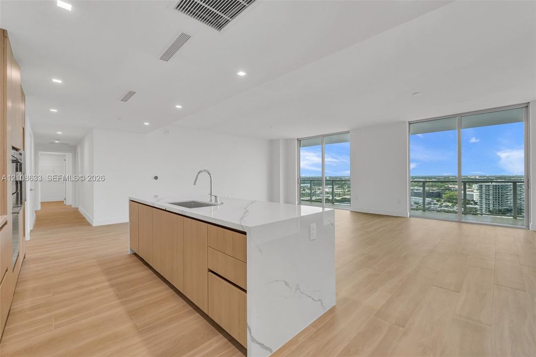 Open-concept kitchen featuring an island with a waterfall-edge countertop, light wood-finish cabinetry, wood-finish flooring, and floor-to-ceiling glass sliding doors