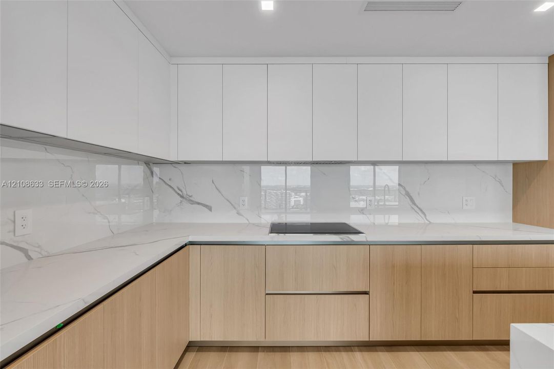 Contemporary kitchen featuring flat-panel upper cabinetry, light-toned wood base cabinetry, stone-surface countertops, full-height stone-surface backsplash, and a built-in cooktop
