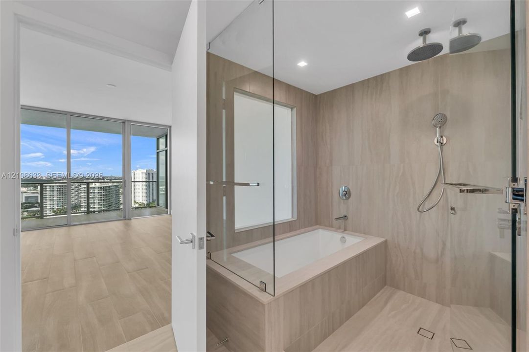 Bathroom featuring floor-to-ceiling large-format tile, glass-enclosed wet room with built-in soaking tub, dual rainfall shower heads, and hand-held shower fixture