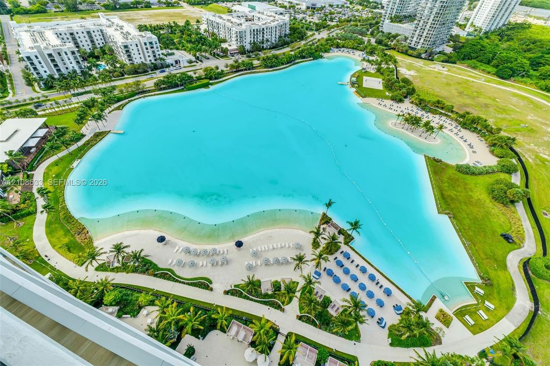 Large-scale turquoise lagoon featuring sandy shorelines, paved pedestrian pathways, manicured green space, and perimeter palm trees