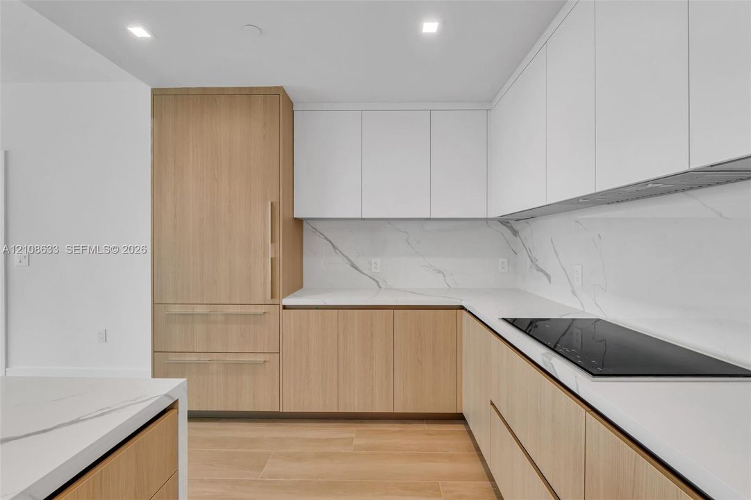 Modern kitchen featuring flat-panel wood-finish cabinetry, floor-to-ceiling refrigerator paneling, white solid-surface countertops, stone-patterned backsplash, and an integrated induction cooktop