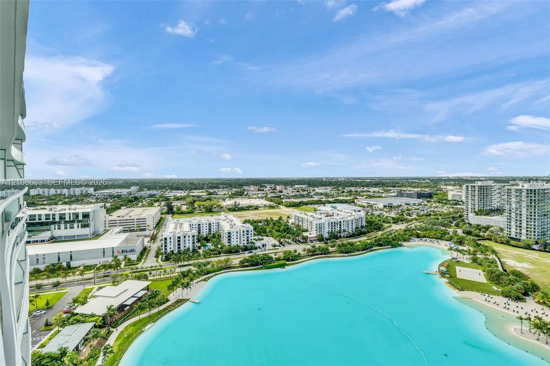 Elevated balcony featuring a glass panel railing, showcasing a panoramic perspective of a large turquoise lagoon, sandy beach area, and surrounding landscape