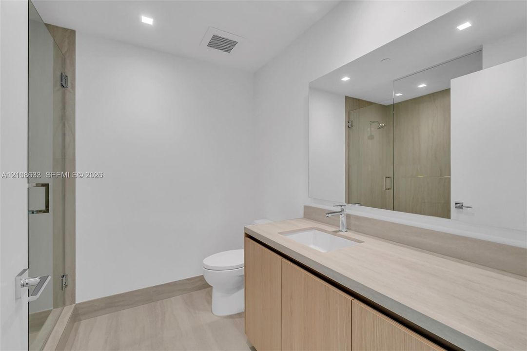 Floating vanity with light wood-finish cabinetry, integrated rectangular sink, large wall-spanning mirror, and glass-enclosed shower with neutral-toned wall tiling