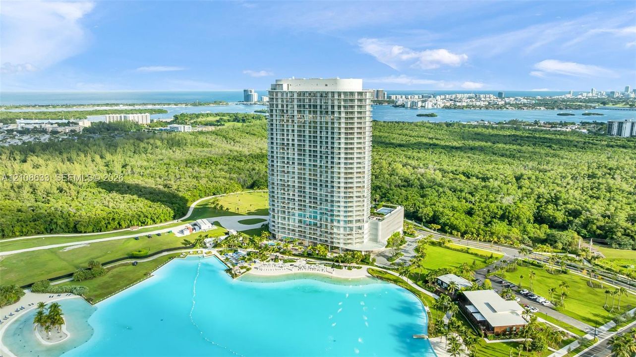 High-rise residential tower featuring floor-to-ceiling glass windows, wraparound balconies, an expansive crystalline lagoon, landscaped grounds, and paved parking areas