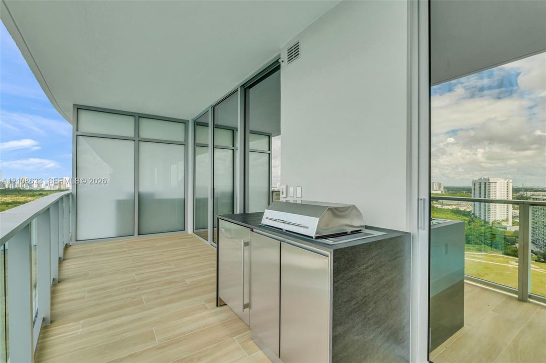 Covered balcony featuring wood-finish tile flooring, glass railings, and a built-in outdoor kitchen with stainless steel cabinetry and a metal grill