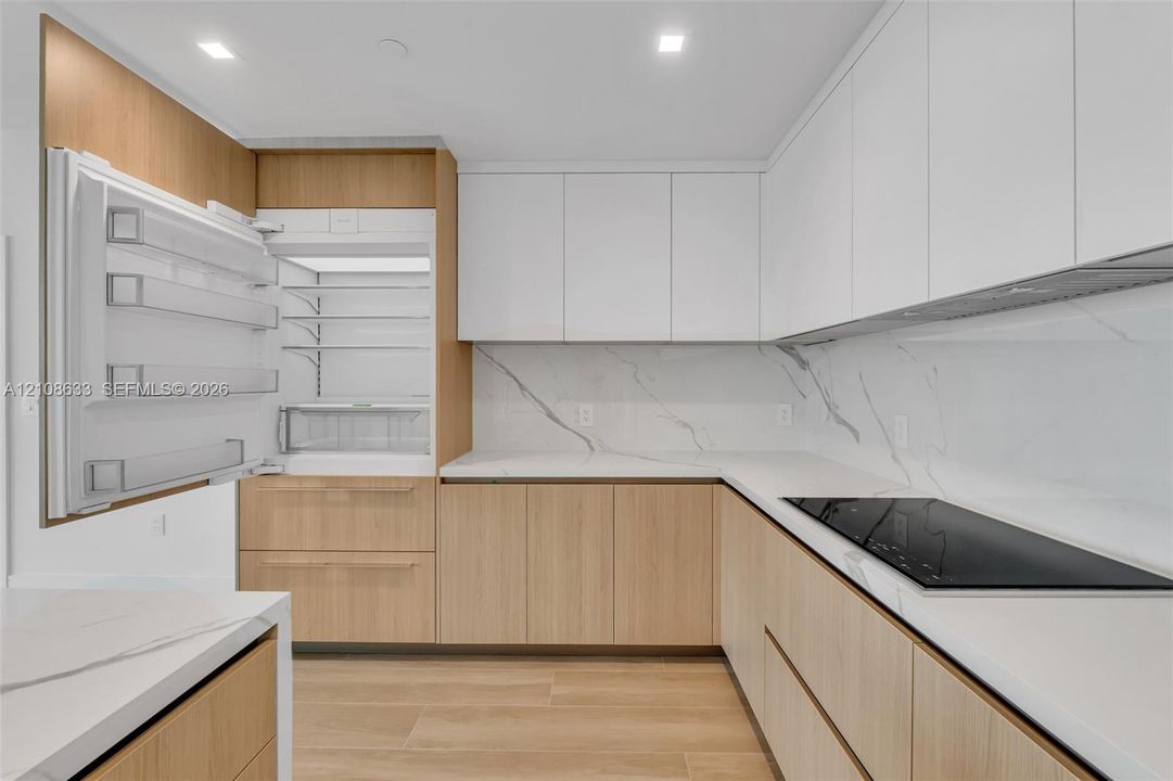 Contemporary kitchen featuring flat-panel wood-finish cabinetry, white matte upper cabinetry, white stone-patterned backsplash, integrated panel-ready refrigerator, and built-in electric cooktop