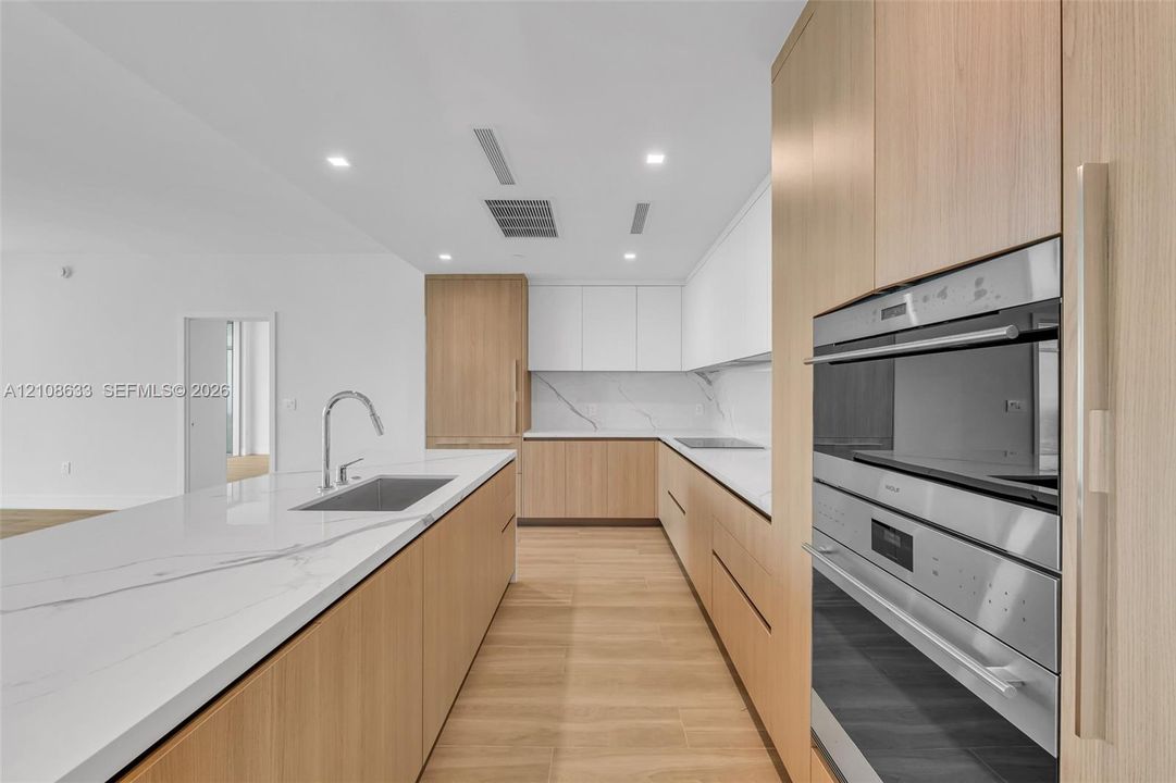 Kitchen featuring wood-finish cabinetry, white stone-look countertops with veining, a stainless steel undermount sink with a gooseneck faucet, and integrated stainless steel wall ovens