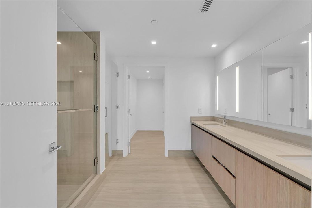 Floating wood-finish vanity with dual integrated sinks, light-toned countertop, floor-to-ceiling glass enclosed shower with integrated niche, and full-width mirror with integrated vertical lighting