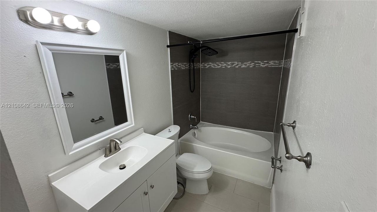 Bathroom featuring a white vanity cabinet with an integrated basin, a wall-mounted light fixture, and a soaking tub surrounded by dark tile with a decorative accent strip
