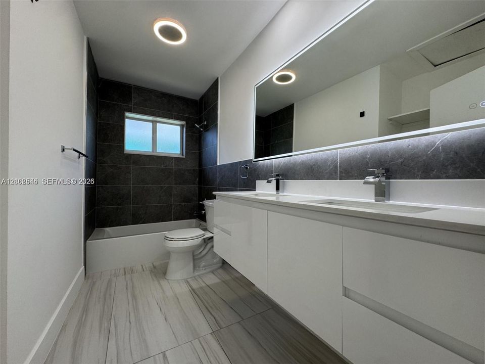 Dual-sink floating vanity with sleek white cabinetry, large illuminated mirror, contrasting dark stone-finish shower wall tile, and light-toned wood-finish flooring