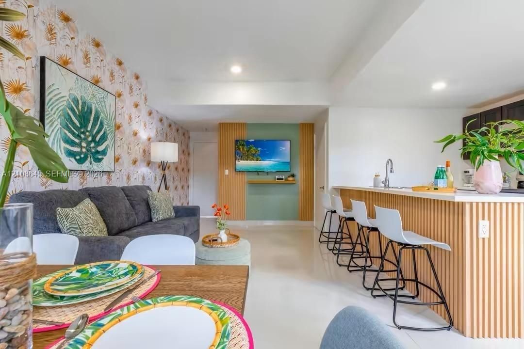 Open-concept interior featuring light-toned tile flooring, custom slatted wood kitchen peninsula, decorative accent wall with patterned wall covering, and a recessed entertainment niche with integrated shelving