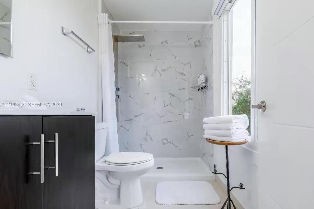 Bathroom featuring a dark wood-finish vanity with a white countertop, a walk-in shower with stone-patterned tile surround, a built-in rainfall showerhead, and a tall rectangular window