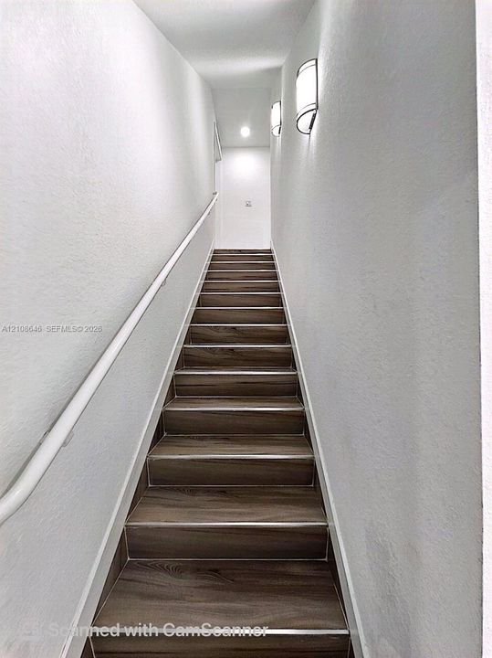 Staircase featuring wood-finish treads, a white handrail, and two wall-mounted curved light fixtures