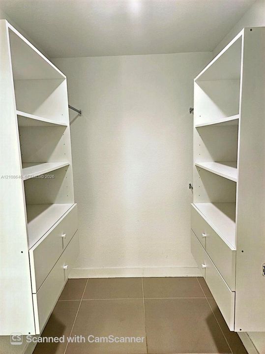 Walk-in storage space featuring custom white cabinetry with integrated shelving, dual lower drawers, and metal garment rods, finished with neutral tile flooring