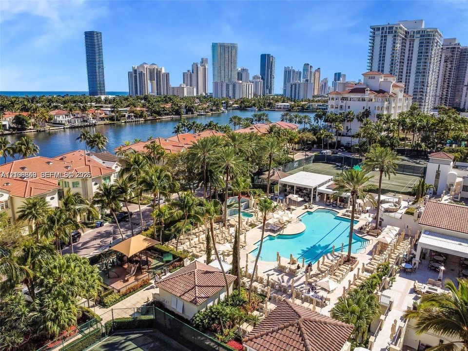 Waterfront community featuring an expansive swimming pool, professional-grade tennis courts, mature tropical landscaping, and tile-roof architecture