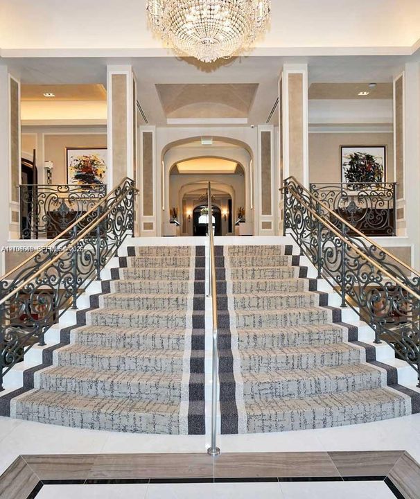 Grand dual staircase featuring intricate wrought iron railings, stone flooring with decorative inlay, and a central crystal chandelier under vaulted ceiling detailing