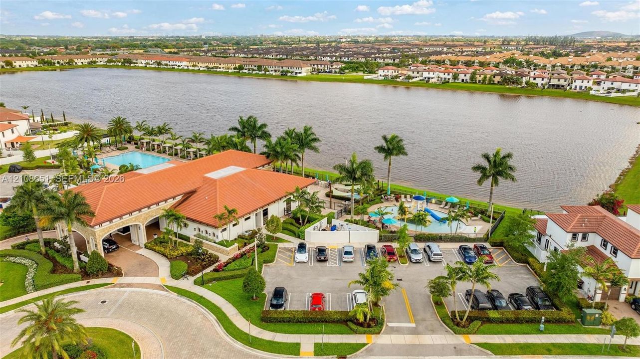 Waterfront clubhouse featuring a tile roof, an outdoor swimming pool, paved parking, and manicured landscaping with palm trees