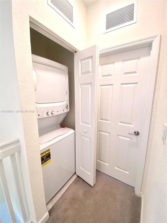 Stackable laundry appliance set housed in a recessed closet with a paneled door, featuring neutral wall finishes and carpeted flooring