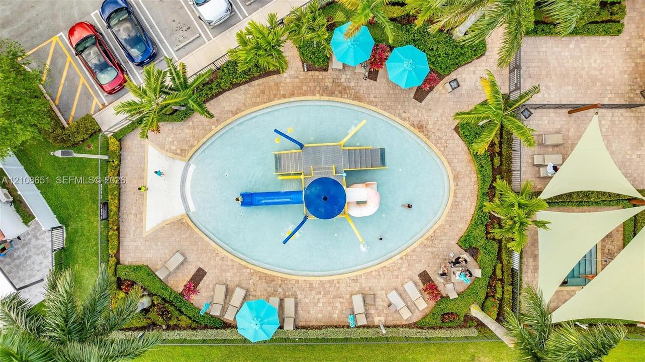 Oval-shaped swimming pool with a central play structure, integrated slide, paver stone pool deck, and landscaped surroundings