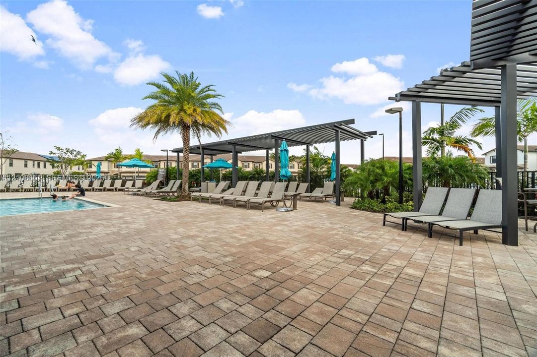 Community swimming pool featuring paved decking, metal pergola structures, and mature palm trees