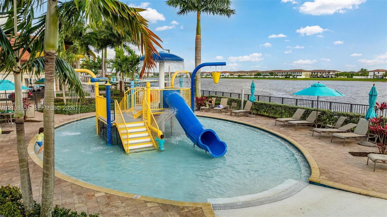 Waterfront community recreation area featuring a shallow-entry pool, integrated water slide, splash pad structure, and stone paver surround