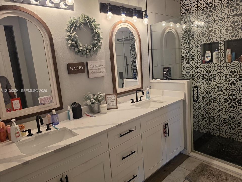 Dual-vanity cabinet with quartz countertops, integrated rectangular sinks, matte black fixtures, arched wood-framed mirrors, and a glass-enclosed shower featuring decorative patterned tilework