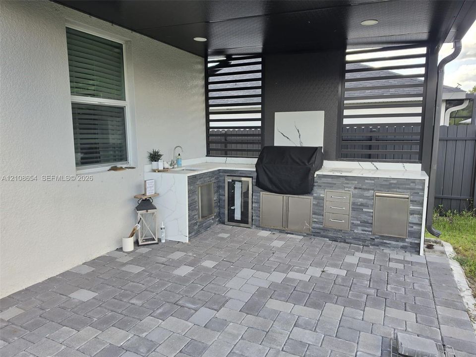 Covered outdoor kitchen featuring stone veneer cabinetry, white veined solid-surface countertops, stainless steel storage, and stone paver flooring under a dark-toned ceiling