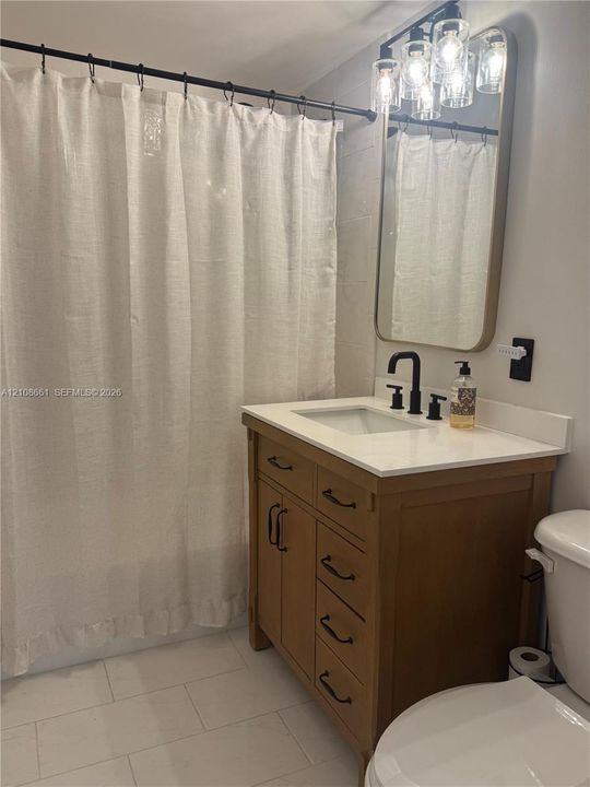Wood-finish vanity with a rectangular sink, stone-composite countertop, matte black fixtures, and a framed mirror with overhead multi-light vanity lighting