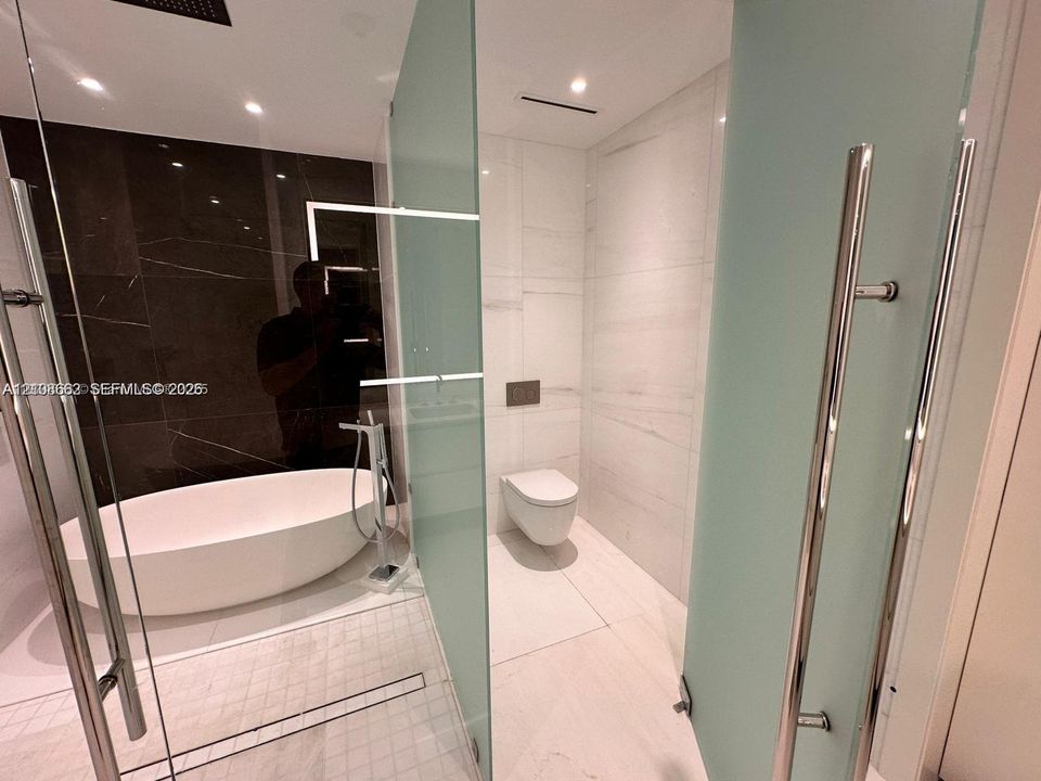 Freestanding soaking tub, floor-to-ceiling dark stone-finish wall tile, frosted glass partitions, wall-mounted toilet, and large-format porcelain tile flooring