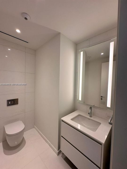 Modern bathroom featuring a wall-mounted toilet, floating vanity with a stone-finish countertop, integrated under-mount sink, chrome fixtures, and a vertical LED-backlit mirror