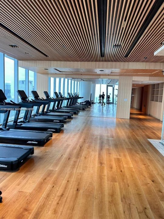 Fitness facility featuring linear wood-slat ceiling detailing, floor-to-ceiling windows, wood-finish flooring, and built-in wall shelving