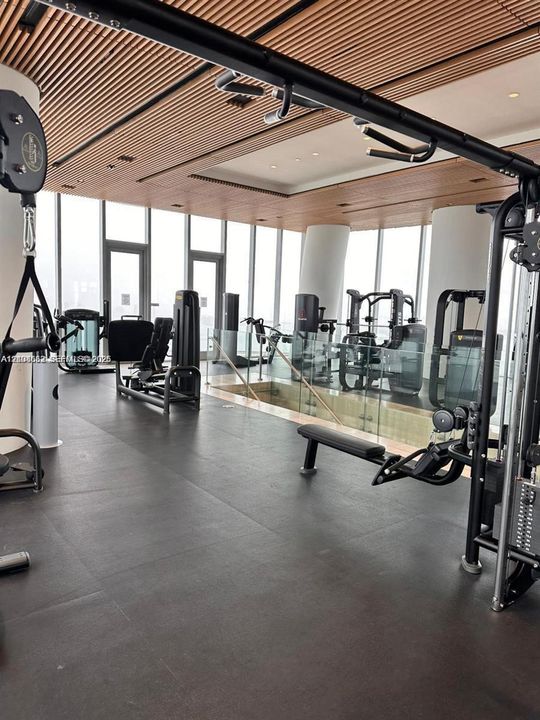 Fitness center featuring floor-to-ceiling windows, linear wood-slat ceiling, dark resilient flooring, and glass partition railings
