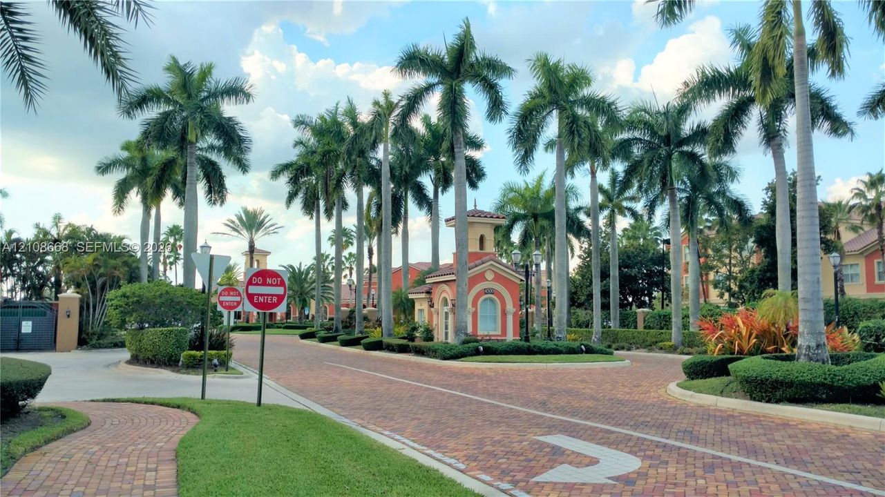 Gated community entrance featuring paver-stone roadways, established palm tree landscaping, and a Mediterranean-style guard pavilion