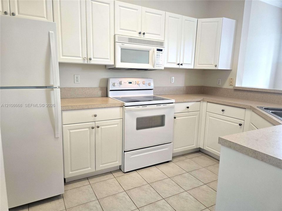 Kitchen featuring white shaker-style cabinetry, neutral-toned countertops, tile flooring, and integrated white appliances including a range and microwave