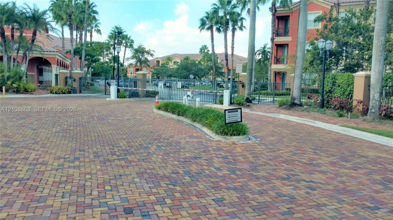 Gated community entry featuring paver-stone driveway, metal security gate, and mature palm trees