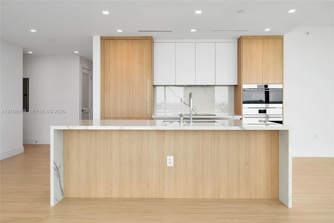 Kitchen featuring a waterfall-edge island, wood-finish cabinetry, white upper cabinets, stone-surfaced countertops and backsplash, and built-in stainless steel appliances