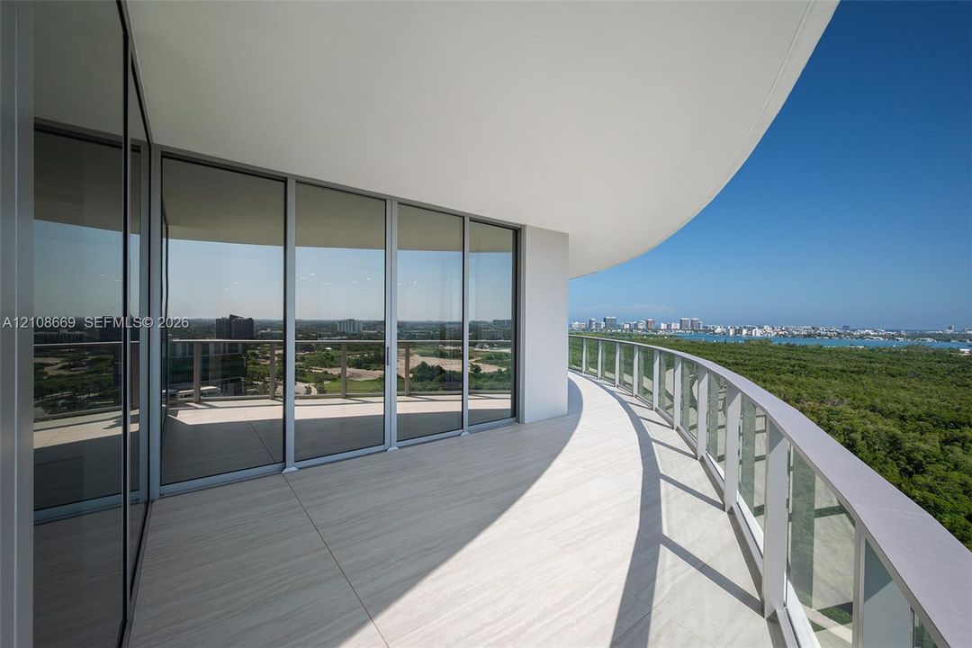 Curved balcony featuring light-toned tile flooring, glass panel railing, floor-to-ceiling sliding glass doors, and a deep overhang