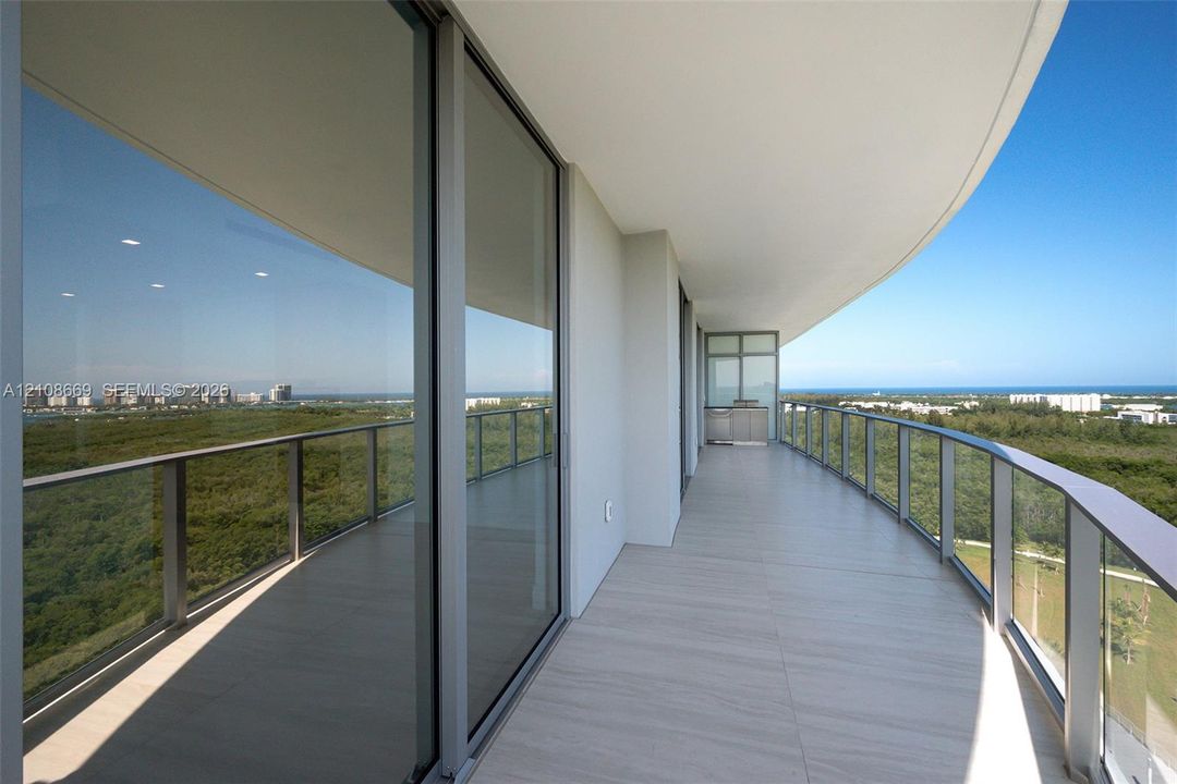Curved covered balcony featuring large-format tile flooring, a glass-panel railing, floor-to-ceiling sliding glass doors, and a built-in outdoor kitchen station