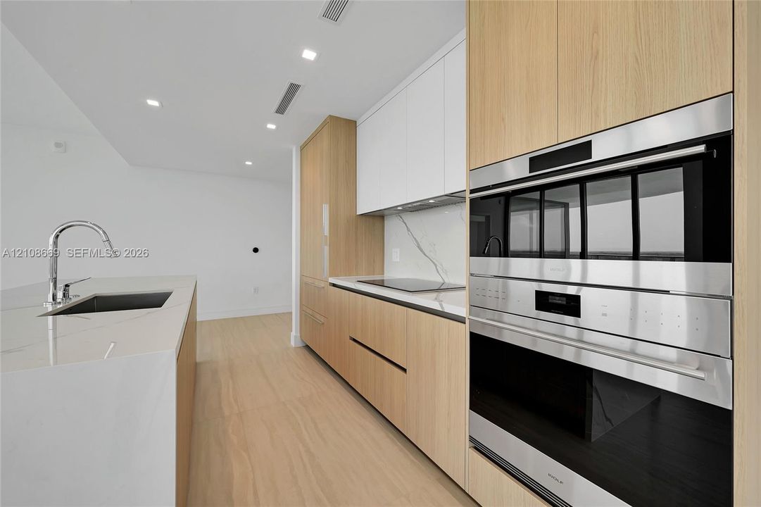 Kitchen featuring light wood-finish cabinetry, quartz countertops with a veined pattern, integrated stainless steel double wall ovens, a sleek electric cooktop, and a kitchen island with an undermount sink and chrome faucet