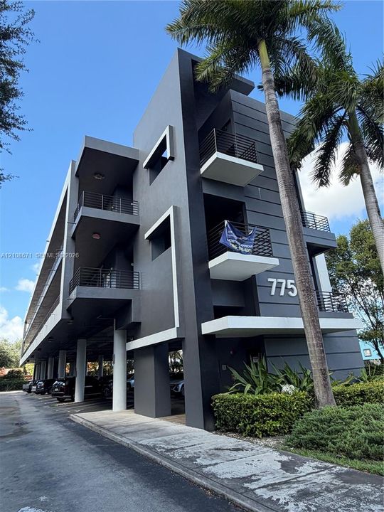 Modern multi-story exterior featuring a gray stucco facade with white geometric accents, stacked private balconies with horizontal metal railings, and covered ground-level parking