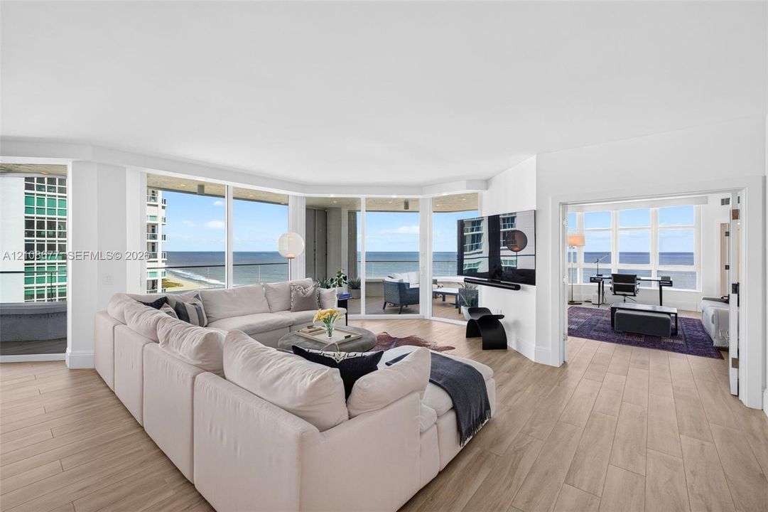 Expansive living area (and bedroom to the right, currently being used as a den) w/ hurricane impact rated floor-to-ceiling windows, wood look plank porcelain flooring, recessed lighting, and a wrap-around balcony with panoramic ocean views. Light filtering remote operated shades with Bandalux