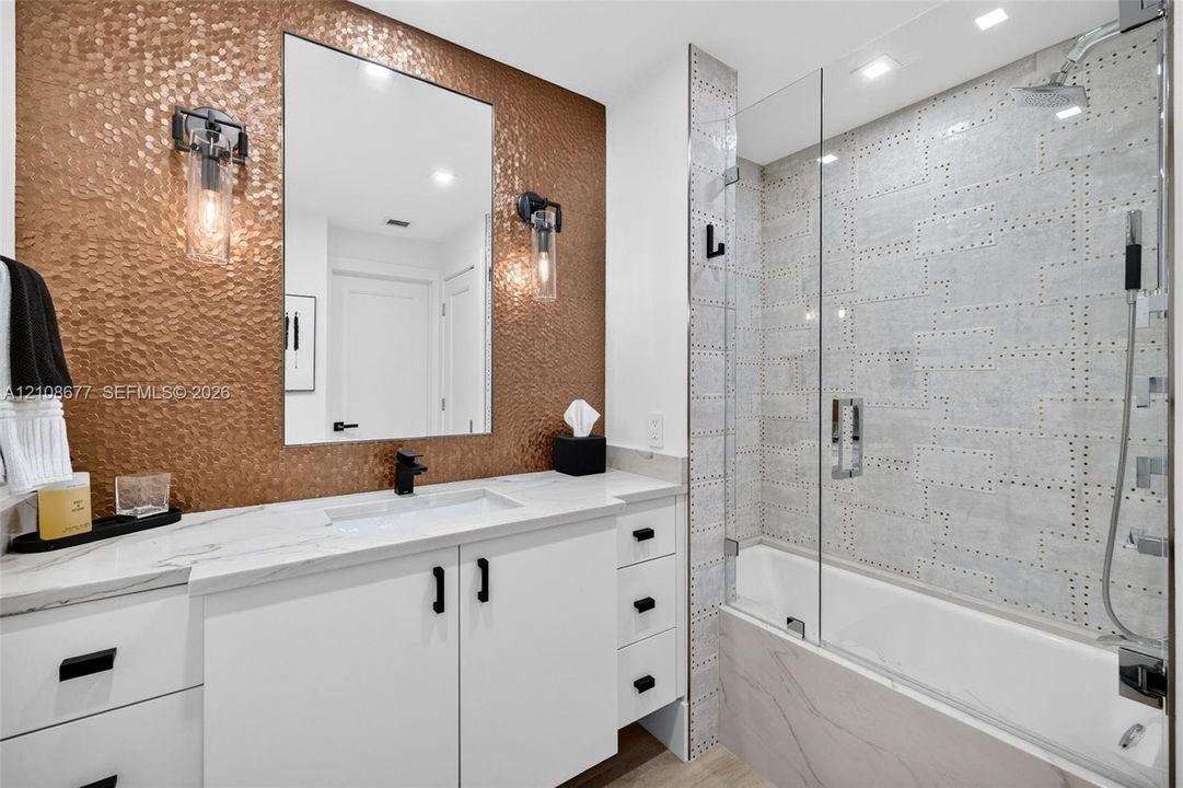 Bathroom featuring a hammered metallic tile accent wall, white floating vanity with stone counters, undermount sink, frameless glass shower enclosure, and custom decorative wall tile. Tub / shower combo w/ handheld shower head & rain head