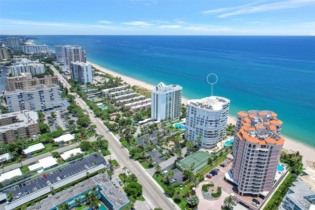 Aerial perspective of a multi-story residential building located on the coastline, featuring an on-site tennis court, swimming pool, and direct proximity to the ocean and sandy beach