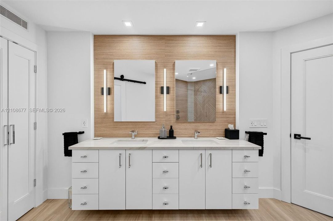 Dual-sink custom vanity featuring white cabinetry, light-toned stone counter, warm wood inspired wall tile and wall-mounted vertical sconces