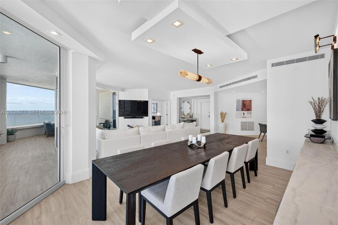 Open-concept interior featuring wood look plank porcelain flooring, recessed lighting, a custom architectural ceiling, a modern linear chandelier, and floor-to-ceiling glass doors opening to an exterior balcony from dining area w/ built in custom buffet for extra storage. Sliders lead again to the balcony for entertaining