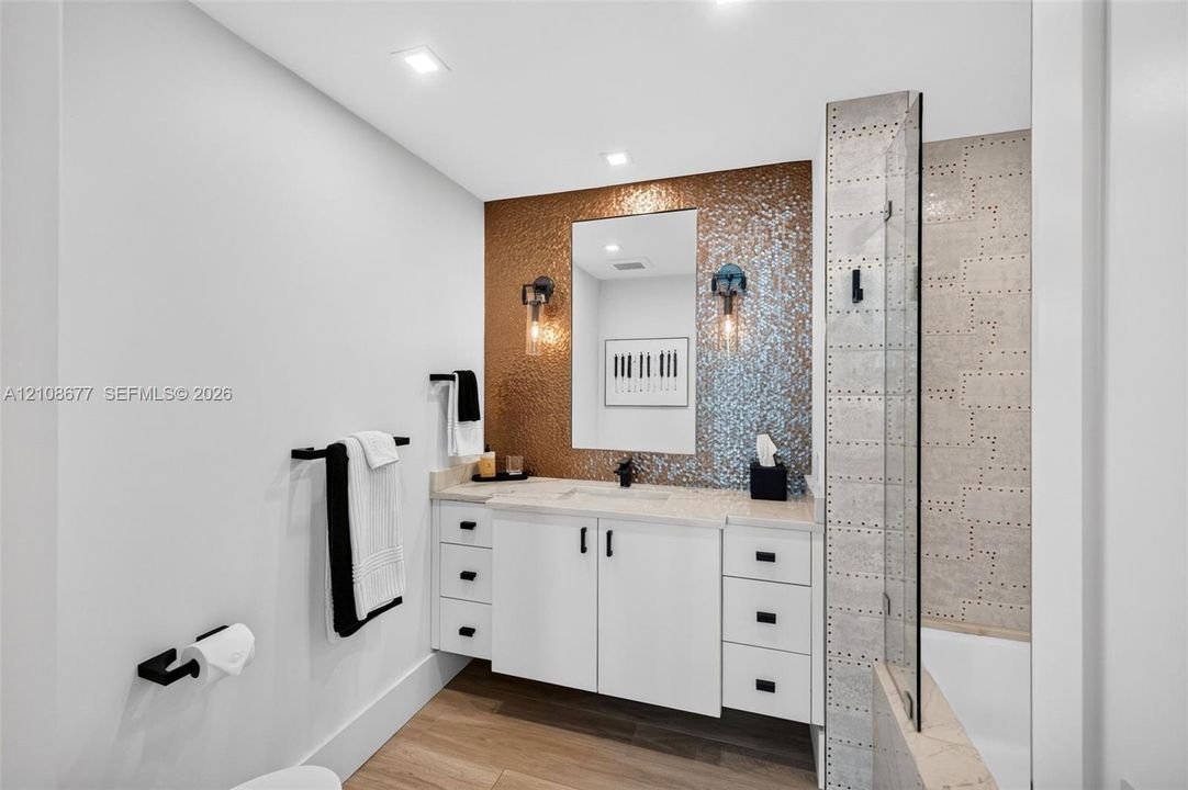 Bathroom w/ custom wall hung white vanity with matte black hardware, metallic mosaic tile accent wall, recessed lighting, and a glass-enclosed shower with custom industrial-inspired wall tile