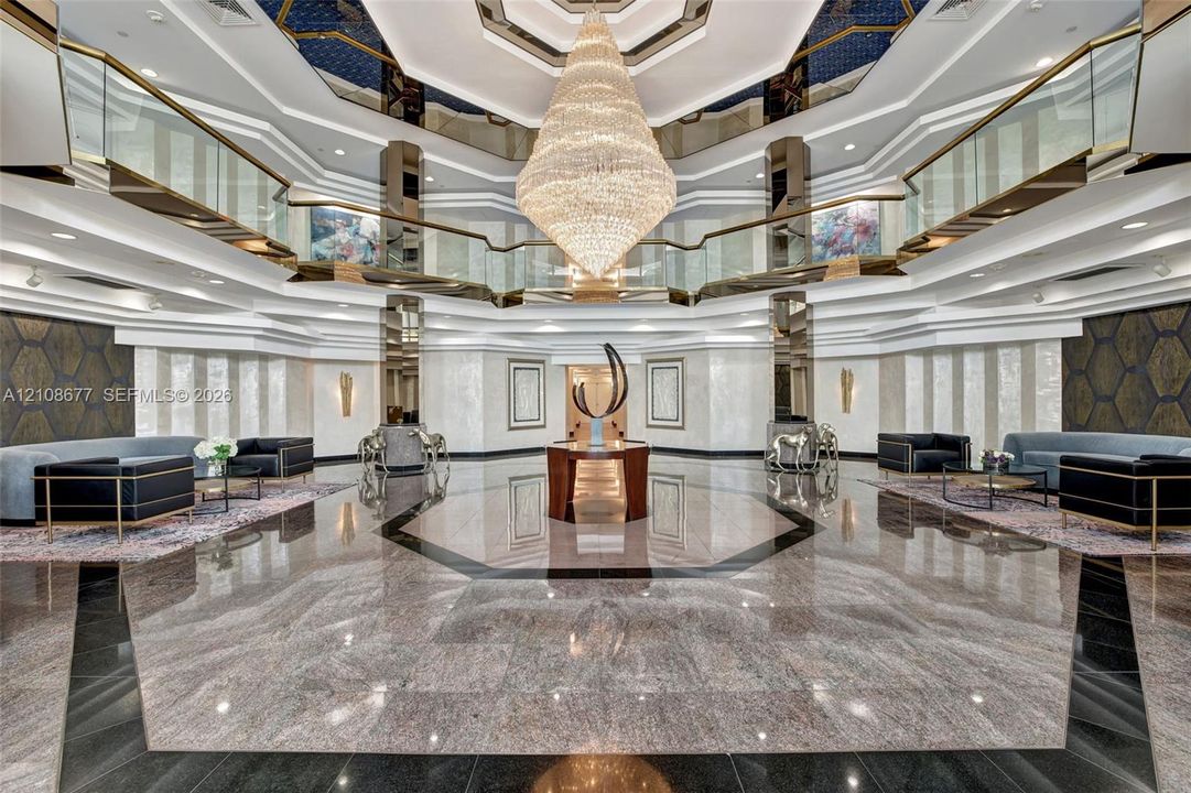 Grand lobby foyer featuring an expansive polished stone floor, a suspended multi-tiered crystal chandelier, a multi-faceted coffered ceiling with recessed lighting, and a wrap-around glass-paneled mezzanine and newer lobby furniture