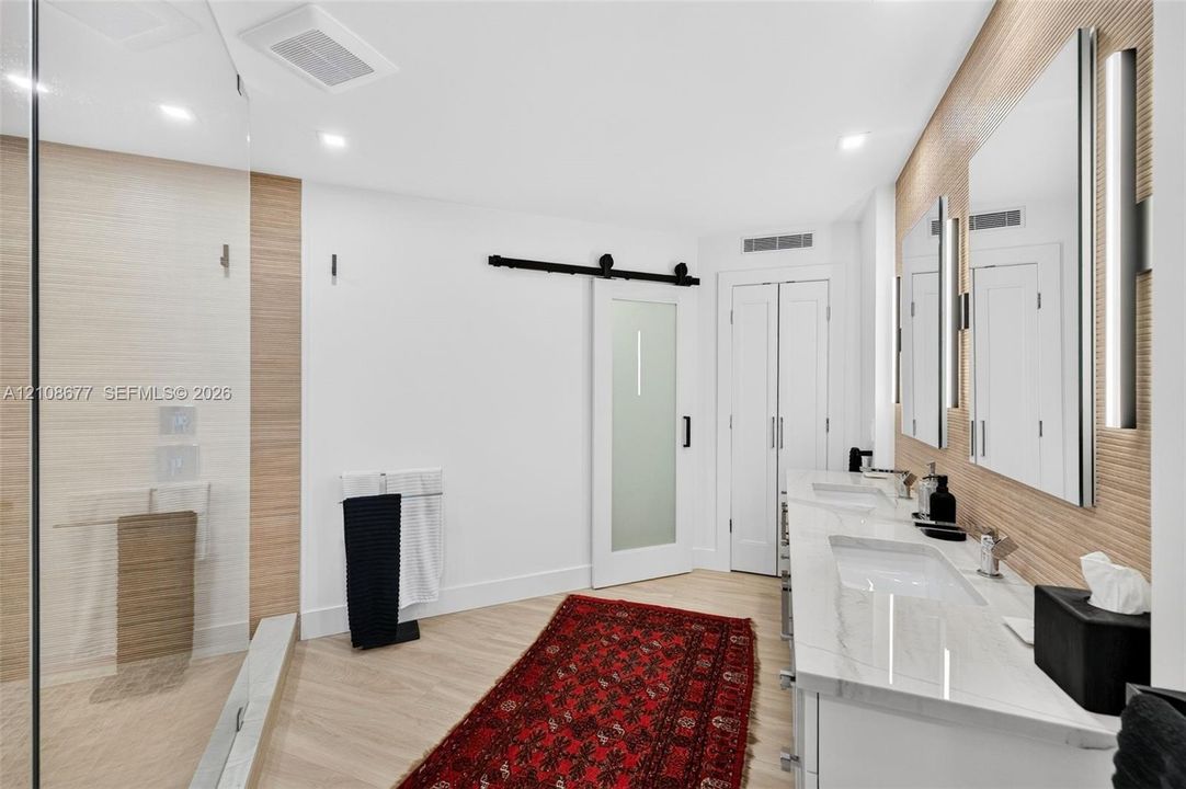 Double vanity with stone-surfaced countertops, frameless glass-enclosed shower, textured wood look wall tile, wall sconces, and frosted glass sliding door to water closet. Tall linen closet next to that.