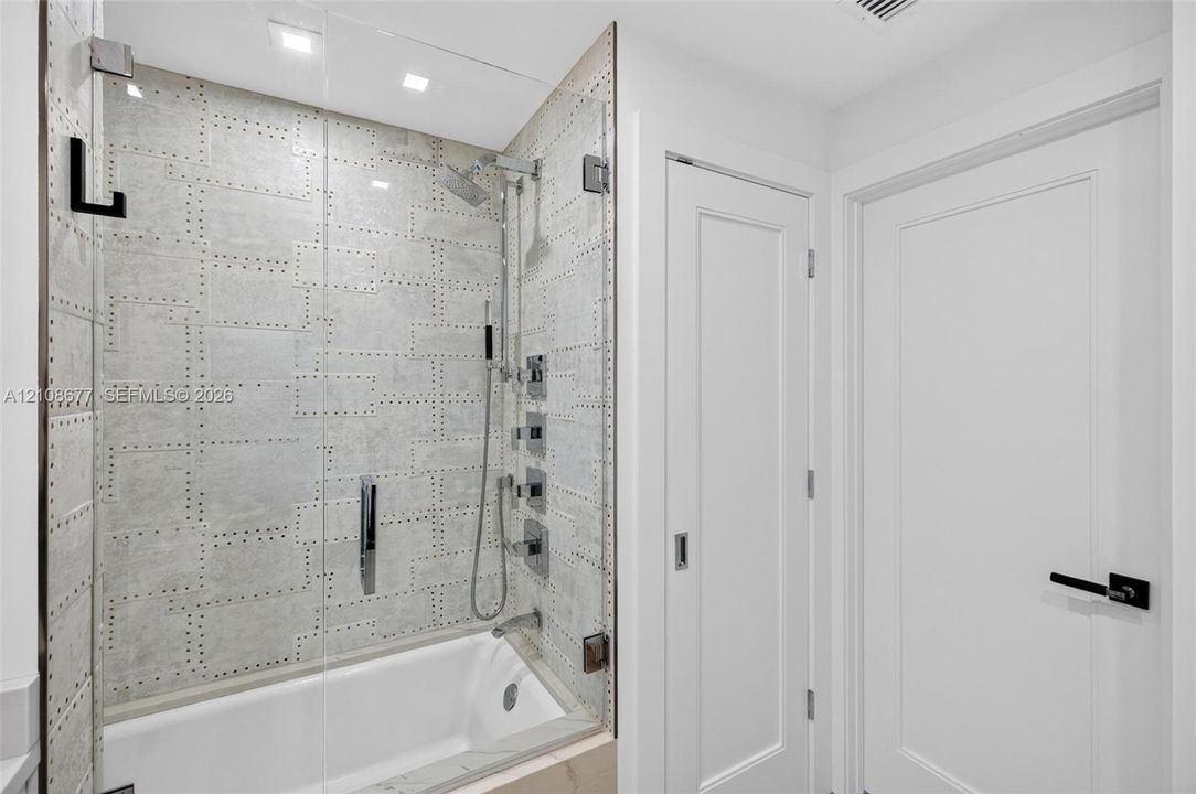 Shower enclosure featuring luxury patterned wall tile, frameless glass door, adjustable-height handheld showerhead, wall-mounted faucet controls, and recessed ceiling lighting plus linen closet
